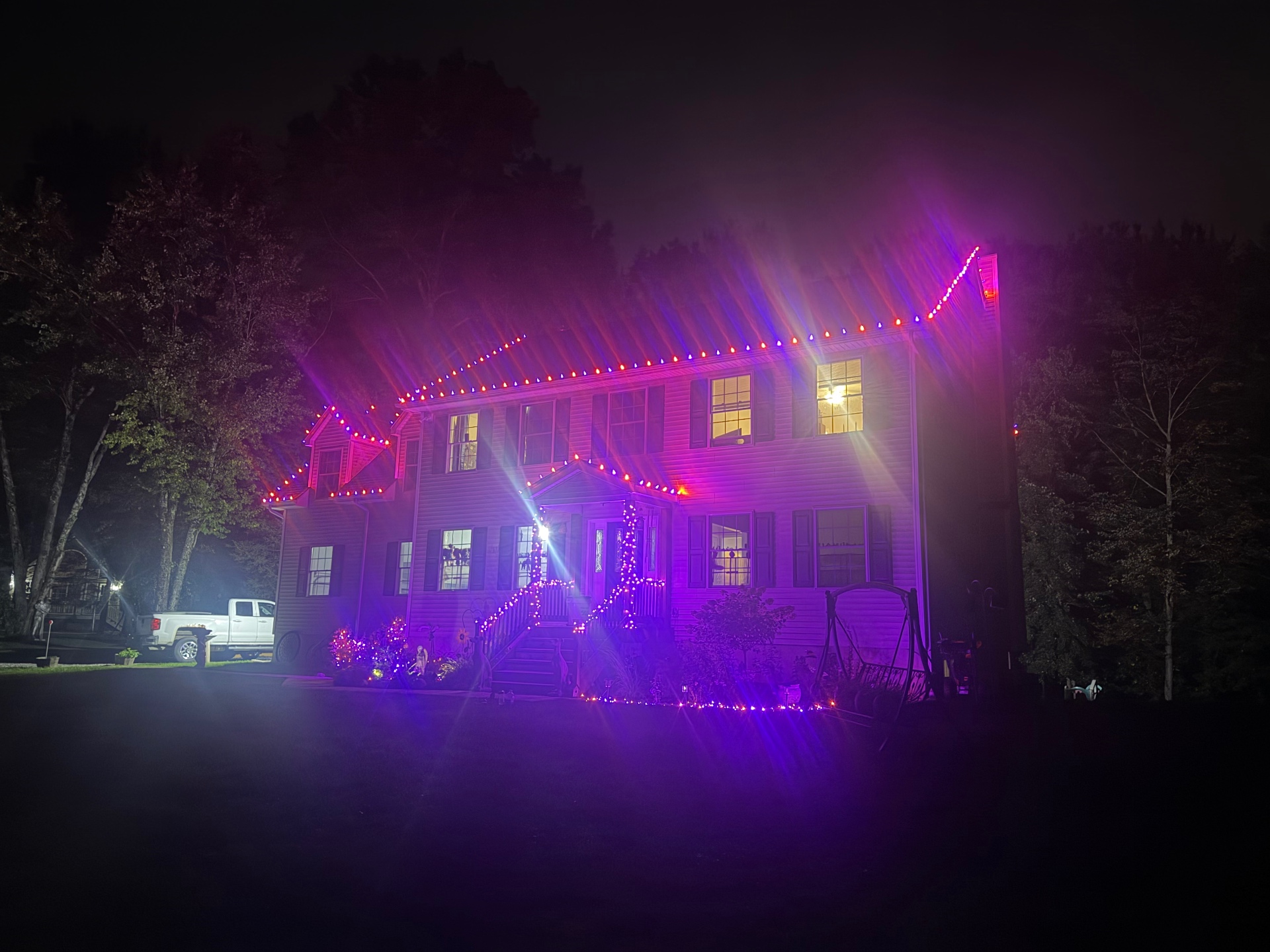 Home with dormer and porch roofline lights glowing beneath mature trees at night