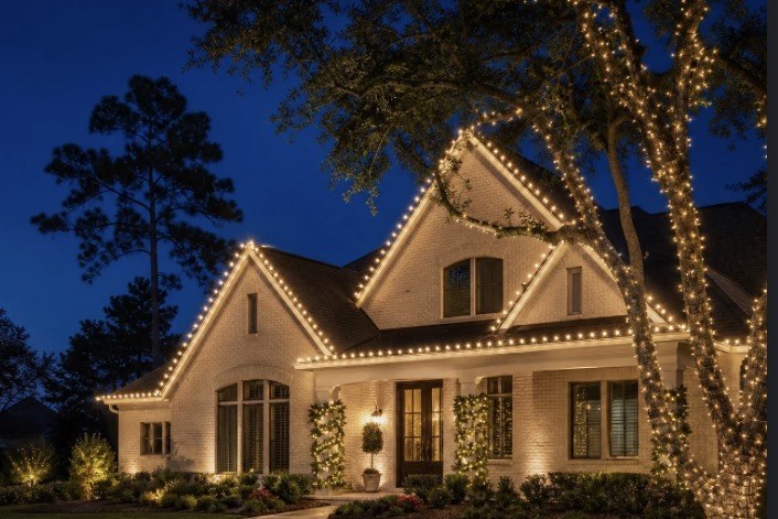 White brick home and mature trees wrapped in warm white holiday lights