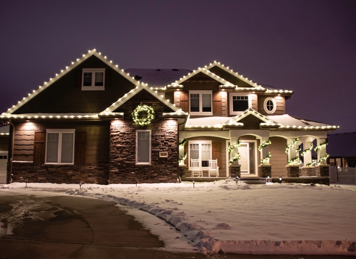 Snowy home with roofline lights, a lit wreath and decorated porch columns